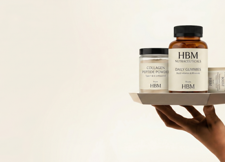 A hand holds a grey tray displaying three HBM Nutraceutical consumer behaviour trends in the supplement industry: a jar of unflavored Collagen Peptide Powder, a large bottle of Daily Gummy Vitamins, and a sachet of Electrolyte Boost. This high-end photo uses minimalist lighting to convey transparency to informed consumers.